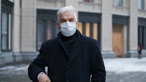 Old retired businessman or gentleman in coronavirus facemask due to lockdown restrictions. Mature adult elderly male in black coat walks on city streets. Senior man with graying hair in covid-19 mask.