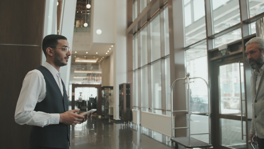 Slowmo shot of young male concierge in uniform welcoming guest couple at hotel lobby, escorting them to reception desk and communicating politely Royalty-Free Stock Footage #1077134351