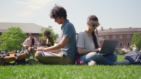 Couple of students sitting in park and studying with laptop and textbook. Portrait of young caucasian man and woman prepare for exam at campus lawn