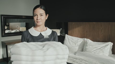 Medium portrait of young female housekeeper smiling to camera while holding fresh white towels standing in luxury hotel room