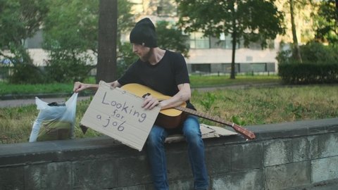 Homeless man with belongings on the street playing guitar, looking for a job. broken down person, gotten into poverty, misery, been laid off, fired, sacked. social problems, unemployment concept