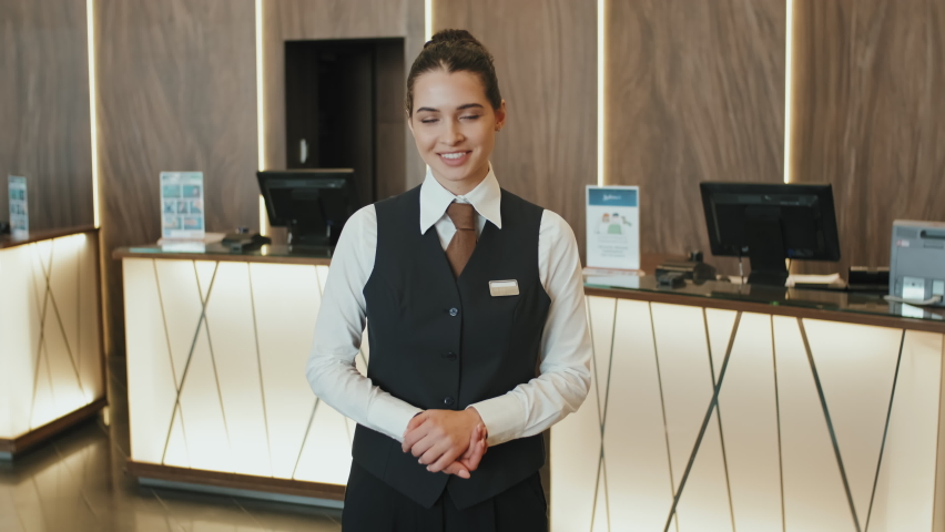 Medium slowmo portrait of young smiling female hotel manager or receptionist in uniform posing for camera standing in modern lobby with counters for check-in at background Royalty-Free Stock Footage #1077142838
