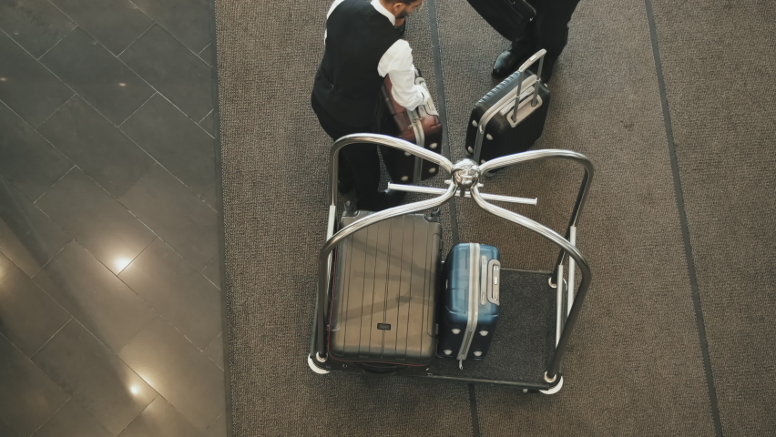 Top-view shot of contemporary tourist couple giving their suitcases to young male porter placing them on luggage cart in high-class hotel lobby Royalty-Free Stock Footage #1077145457