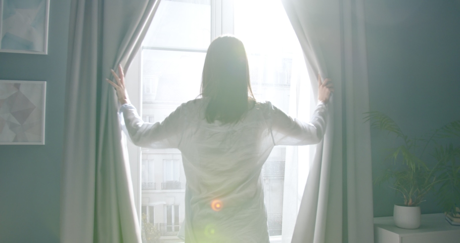 Back view of Caucasian young rested woman waking up after sleeping stands up from bed and opening curtains looking at window at home in sunny morning. Female awake after healthy sleep greeting new day