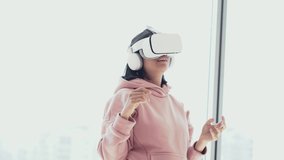 The girl is having fun in virtual reality glasses, stands against the background of the window talking to someone and showing the class. High quality 4k footage