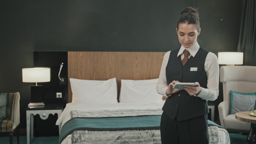Medium slowmo portrait of young female hotel manager in uniform smiling to camera with digital tablet in hands standing in modern comfortable hotel room Royalty-Free Stock Footage #1077147776