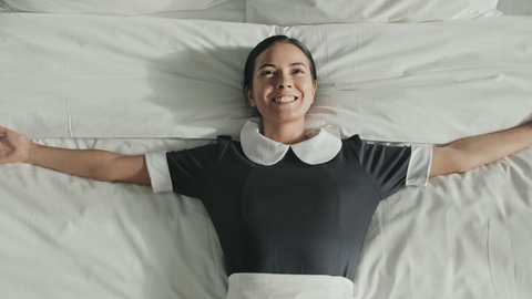 Slowmo shot of relaxed female housekeeper lying down on her back on luxury hotel bed with white linens with arms spread resting after work