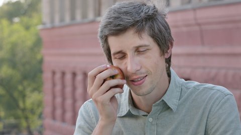 Young male vegetarian vegan holds beautiful ripe apple, inhales delicious aroma. Attractive dieting guy enjoying healthy vitamin breakfast or lunch, sniffing fragrant fruit, closing eyes admiringly