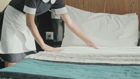 Midsection shot of unrecognizable female housekeeper in black and white uniform making bed while preparing luxury hotel room for guests