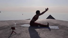 Young sporty fit woman coach doing yoga outdoors. Female instructor teaching online yoga lessons through video call on her mobile phone at sunrise - Streaming lessons and healthcare lifestyle concept