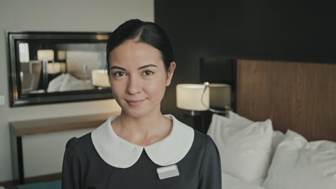 Slow-motion medium close-up portrait of young female housekeeper in uniform smiling to camera standing in modern hotel room