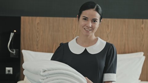Tilting-up portrait with slowmo of young smiling woman working as housekeeper in hotel holding white towels standing next to king-size bed in hotel room and posing for camera