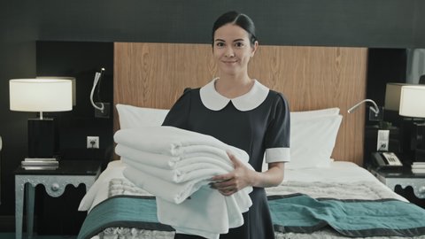 Medium slowmo portrait of young female housekeeper in uniform holding white towels standing next to king-size bed in modern hotel room smiling to camera