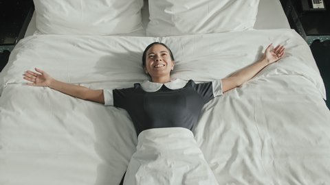 Slowmo shot of young happy female housekeeper in uniform lying down on comfy king-size bed in luxury hotel room with arms spread, enjoying fresh linens
