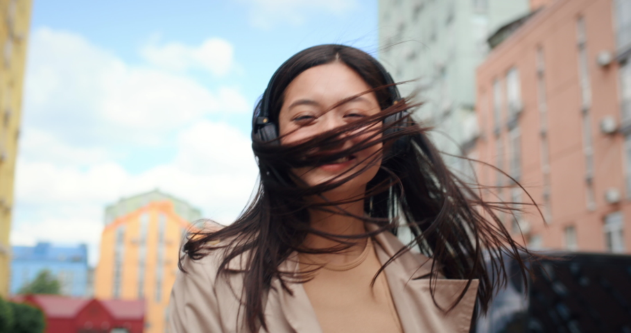 Happy  Asian Girl in Headphones turning to the camera and Smiling. Chinese Girl  Teenager  enjoys listening to  Music using Apps during stroll in City  Royalty-Free Stock Footage #1077151730