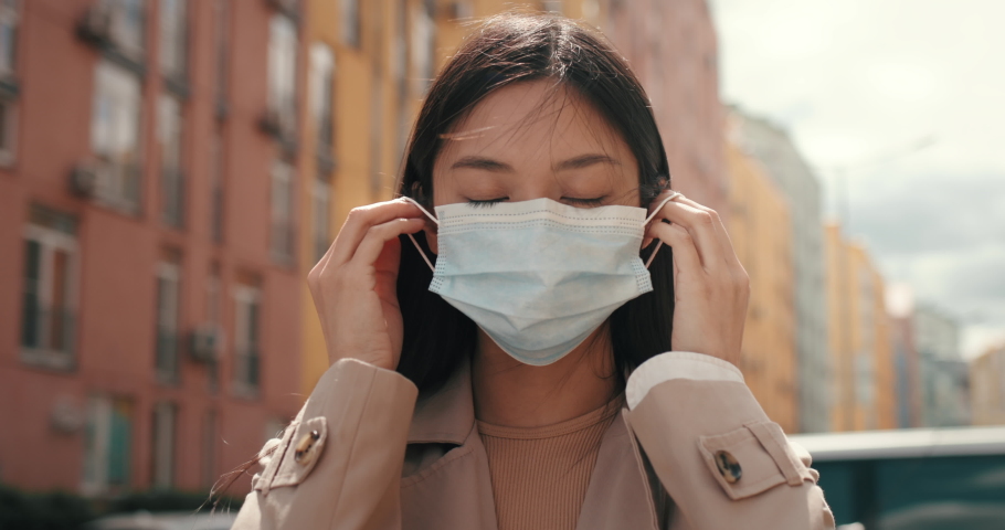 Portrait of Young Sad Asian Girl puts on Protective Mask on Face. Mixed Race Woman with Medical Mask standing outdoors Protects Herself. CoronaVirus outbreak. Pandemic. Asia. Healthcare. Quarantine. Royalty-Free Stock Footage #1077151763