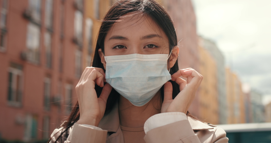 Portrait of Young Sad Asian Girl puts on Protective Mask on Face. Mixed Race Woman with Medical Mask standing outdoors Protects Herself. CoronaVirus outbreak. Pandemic. Asia. Healthcare. Quarantine. | Shutterstock HD Video #1077151763