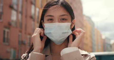 Portrait of Young Sad Asian Girl puts on Protective Mask on Face. Mixed Race Woman with Medical Mask standing outdoors Protects Herself. CoronaVirus outbreak. Pandemic. Asia. Healthcare. Quarantine.