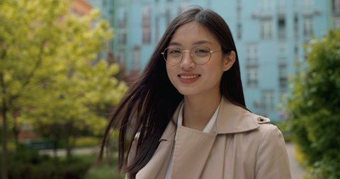 Portrait of Charming Asian Girl in Eyeglasses standing near Colorful Buildings. Pretty Young Chinese Woman looking into Camera and Smiling. Wears Trendy Coat. Attractive Appearance of Ethnic Female.