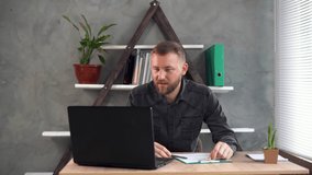 A bearded business man sits at his desk at home in his office, makes a video call, shows the client a tablet with completed forms and tells something.