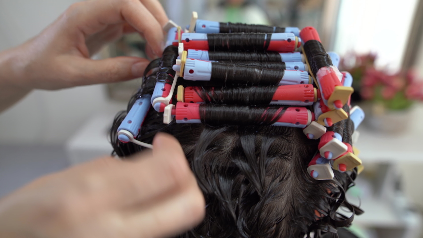 Close-up of a woman from behind with curlers on her hair while a perm, the master removes the curlers from her hair. Royalty-Free Stock Footage #1077158309