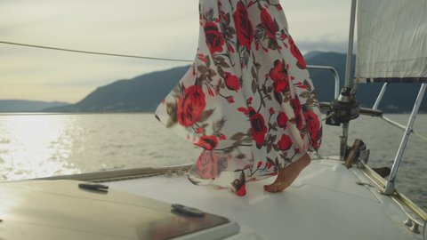 Cropped view of legs, woman standing on yacht bow. Dress with flowers fluttering in wind. Summer vacation at sea, sailing and travelling concept.