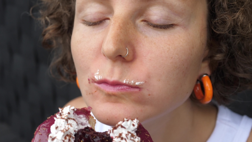 Close up of a young female taking a bite of vegan berry doughnut and chews it with face covered in frosting with her eyes closed 