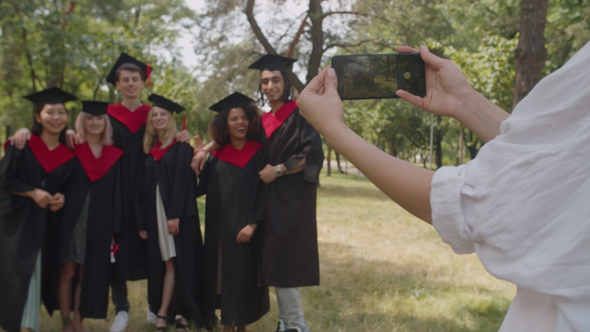 Close-up of female hands taking photo of blurry carefree diverse multiracial graduates in graduation gowns and mortarboards on smart phone outdoors after graduation ceremony. Focus on phone screen. | Shutterstock HD Video #1077165284