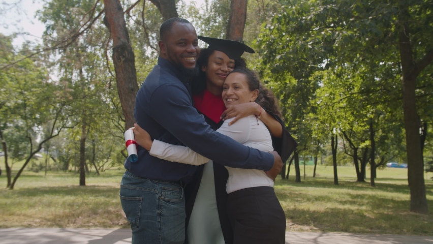 Joyful proud multiracial parents embracing cheerful pretty mixed race female graduate with diploma,dressed in graduation gown and mortarboard, expressing love, care and happiness at graduation day. | Shutterstock HD Video #1077165296
