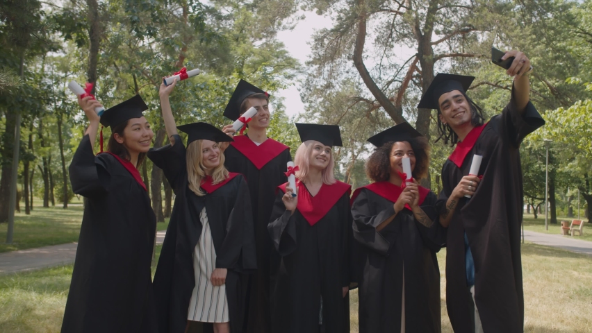 Successful carefree attractive diverse multiracial graduates in graduation gowns and mortarboards holding diplomas with pride, posing for selfie shot on cellphone after outdoor graduation ceremony.