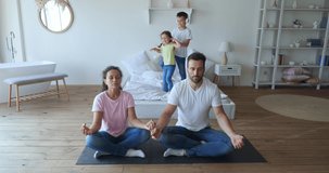 Serene parents meditating with calm face in bedroom while their kids screaming, jumping, dancing and shouting behind at home, family concept, yoga meditation. 4 K video footage