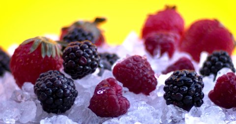 Raspberries, strawberries and blackberries on cold ice on a yellow background. A drop of water falls on the blackberry. Natural cold fruits, juicy and aromatic food.  Slow motion, 8K downscale. 4K.