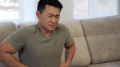 Asian Man feeling stomach pain at home, gastritis symptom, peptic ulcer, pancreatitis. Terrible pain in the stomach. A young man suffering from abdominal pain holding his stomach, sitting on the sofa.