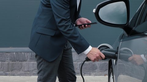 Unrecognizable businessman plugging in charging cable to to electric vehicle. Male hand in jacket inserts power connector into EV car and charges batteries, uses phone for activates start charging