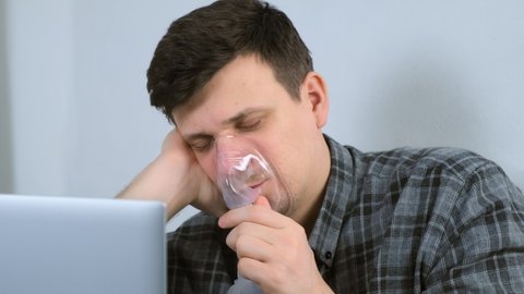 Portrait of sick young man is using nebulizer and inhaler for treatment at home and working on laptop. He is breathing in mask. Fibrosis, covid-19, asthma copd. Treatment, cure and procedure, therapy.