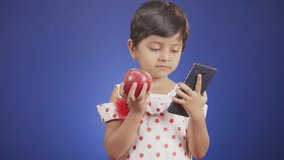 Little girl kid using mobile while eating apple on blue background - concept of childrens mobile addiction and unhealthy distracted lifestyle