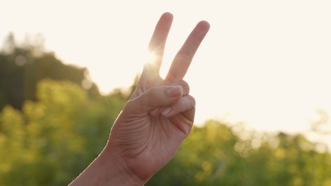 One female hand showing Peace or Victory gesture outdoors isolated on sunny sunset or sunrise sky nature background. Close-up view 4k video footage of hand of woman with two fingers raised up
