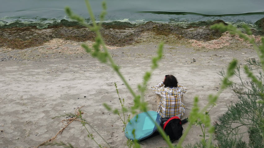 A woman is relaxing on a wild and deserted beach next to her backpack. The camera shoots from above, through the thicket. Waves throw algae onto the shore. The water is green due to blue-green algae.