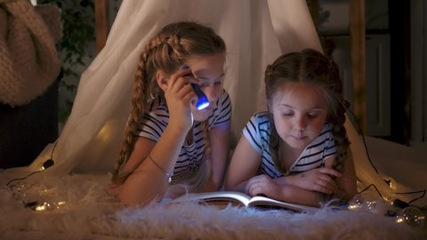 Happy family. Two girl daughters are reading book in tent. Flashlight in hands of girls. Happy family with book in tent. Girl holds. Children lie under the covers and learn to read