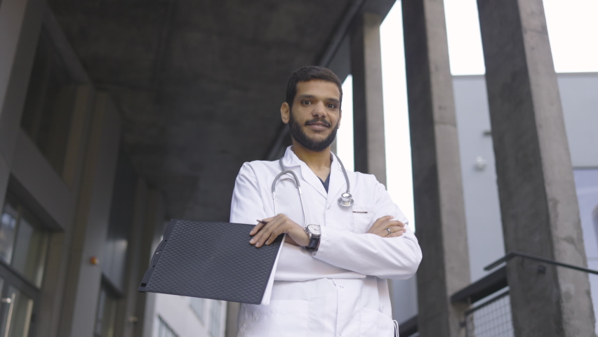 Bottom view of likable confident Arabian male physician in white coat and with stethoscope, holding clipboard and pen, standing with arms crossed on the background of modern hospital. Slow motion, 4K | Shutterstock HD Video #1077185030