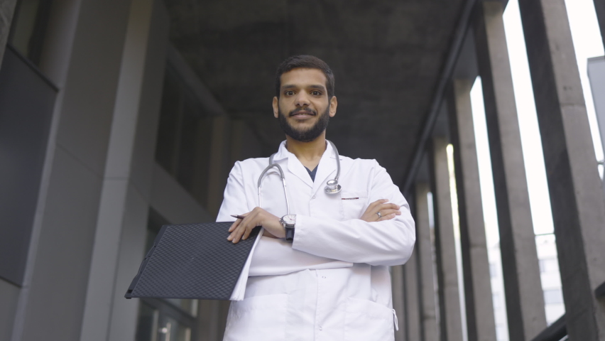 Bottom view of likable confident Arabian male physician in white coat and with stethoscope, holding clipboard and pen, standing with arms crossed on the background of modern hospital. Slow motion, 4K