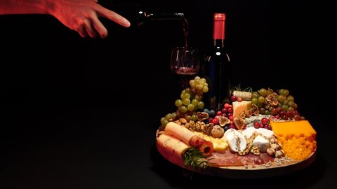 Cheese platter and red wine. Pouring red wine into glass standing on a cheese board with different kinds of cheese. Nicely served selection of exquisite cheese on a black background and wine tasting