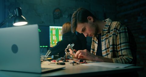 Man Solders in Electronics Workshop. Hacking Attack or IT Startup Concept. Young People Working on Technology Project in Dark Garage or Loft Office. 4K Medium Hand held shot