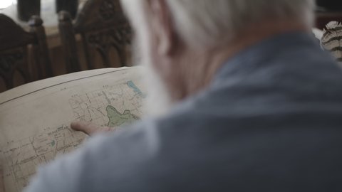 Back View Of An Old Bearded Man Sitting And Holding A Map Pointing Finger On It. View Inside His House In Chester, Idaho, USA. - Selective Focus