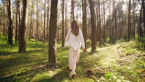 Happy young cheerful woman running away from camera outdoors in pine forest rear view slow motion. Lifestyle Girl walks in park connecting with nature shining in sun at sunset. enjoys life. Happiness