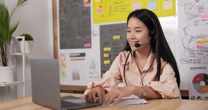 Side view of Smiling asian woman with headset using laptop, talking, working customer support service operator at home or office. Young female in headphones with microphone sitting at workplace desk.