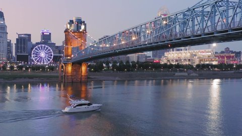 Aerial: Downtown Cincinnati, John A. Roebling Suspension Bridge and motorboat on the Ohio River at sunset. Ohio, USA. 21 September 2019