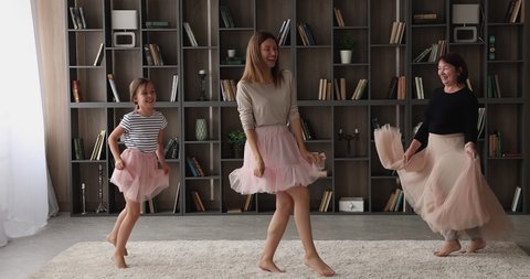 Joyful different generations female family wearing fluffy skirts, dancing barefoot together to energetic funky music in modern living room. Young woman having fun with small daughter and older mother.