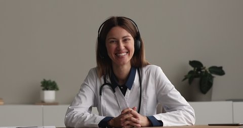 Smiling young professional female gp doctor cardiologist in white coat sitting at table, looking at camera, giving medical healthcare advice, consulting patient distantly in modern clinic office.
