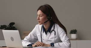 Focused young female doctor general practitioner wearing wireless headset with microphone, giving online video call consultation on computer or taking part in medical educational virtual event.
